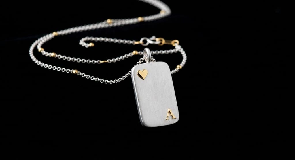 Brushed silver pendant on black: a gold heart at the top left, a gold letter A at the bottom right, and gold beads spaced along the chain.